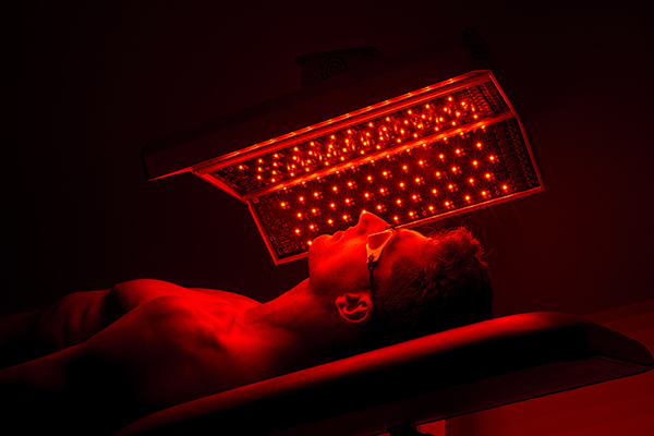 Mann unter rotem LED-Lichtpanel bei einer Lichttherapieanwendung, mit Schutzbrille in dunkler Behandlungsumgebung.