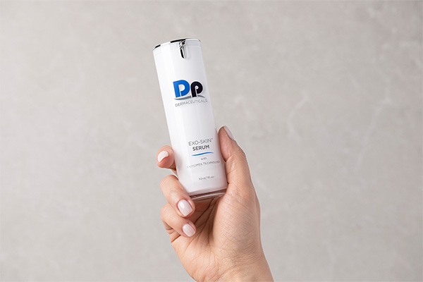 Hand hält ein DP Dermaceuticals EXO-SKIN™ Serum mit weißem Spenderflakon vor hellem, strukturiertem Hintergrund.